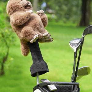 Puede incluir: Una funda para la cabeza del palo de golf de peluche marr&oacute;n con forma de perezoso. El perezoso tiene una expresi&oacute;n sonriente y est&aacute; unido a una funda de punto negra. El palo de golf est&aacute; en una bolsa de golf blanca y negra con otros palos de golf.