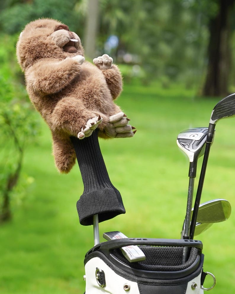 Puede incluir: Una funda para la cabeza del palo de golf de peluche marr&oacute;n con forma de perezoso. El perezoso tiene una expresi&oacute;n sonriente y est&aacute; unido a una funda de punto negra. El palo de golf est&aacute; en una bolsa de golf blanca y negra con otros palos de golf.