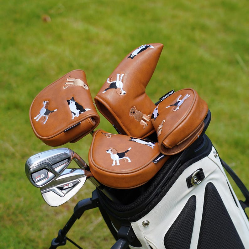 Puede incluir: Una bolsa de palos de golf con fundas de cuero marr&oacute;n con dise&ntilde;os de perros bordados. Los palos de golf son visibles, con la bolsa apoyada sobre una superficie de hierba verde. Las fundas de los palos tienen varias razas de perros.