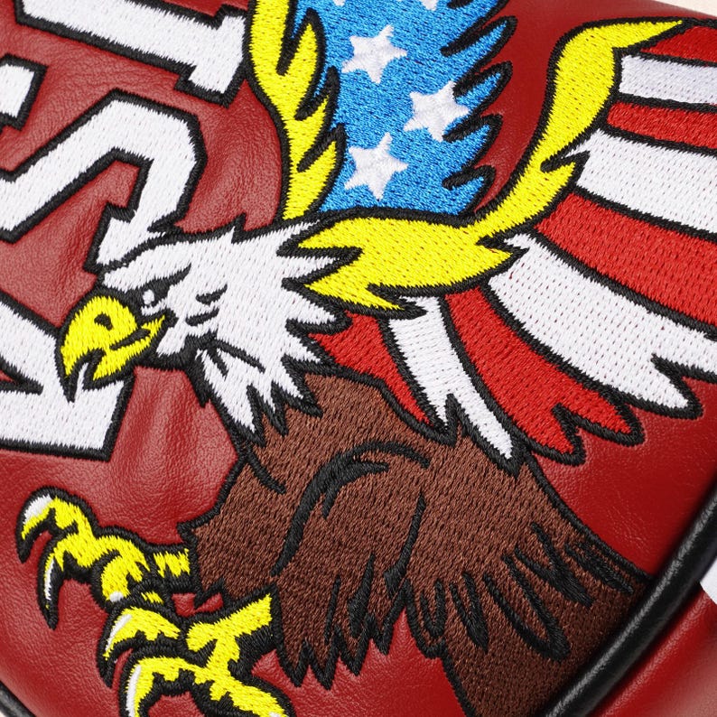 Peut inclure: Gros plan sur un article en cuir rouge avec un motif d'aigle am&eacute;ricain brod&eacute;. L'aigle pr&eacute;sente des couleurs blanches, marron, jaunes, rouges et bleues, avec le drapeau am&eacute;ricain incorpor&eacute; dans les ailes. Des lettres blanches sont visibles.