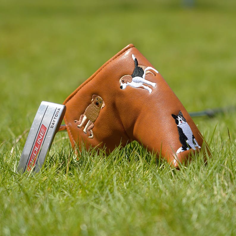 Puede incluir: Una funda de cuero marr&oacute;n para la cabeza del palo de golf con dise&ntilde;os de perros bordados, incluyendo un golden retriever, un beagle y un perro blanco y negro. Un putter plateado con el texto "Daytona" est&aacute; en la hierba.