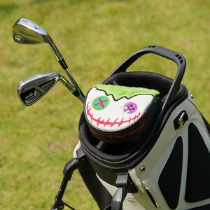 Pode incluir: Uma bolsa de golfe com dois tacos e uma capa de cabe&ccedil;a &uacute;nica. A capa &eacute; branca com um design de rosto costurado, pelo verde e o texto "None of my word" em vermelho. A bolsa &eacute; preta e branca.