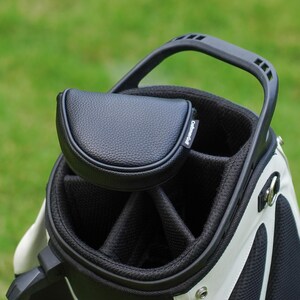 Może przedstawiać: Czarno-biała torba golfowa z pasującą czarną osłoną na putter. Torba ma solidny uchwyt i wiele przegr&oacute;dek. Osłona na putter jest wykonana z teksturowanego czarnego materiału i ma napis "Craftsman Golf".