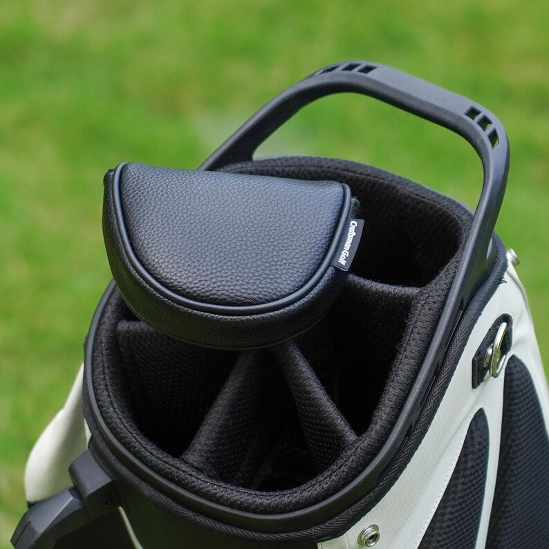 Może przedstawiać: Czarno-biała torba golfowa z pasującą czarną osłoną na putter. Torba ma solidny uchwyt i wiele przegr&oacute;dek. Osłona na putter jest wykonana z teksturowanego czarnego materiału i ma napis "Craftsman Golf".