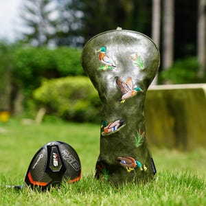 May include: An olive green golf club headcover featuring embroidered mallard ducks and cattails, positioned next to a black and orange TaylorMade M6 golf club driver with a carbon fiber texture on the club head face and top surface.