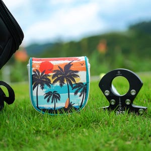 May include: A golf club head cover with a tropical sunset design featuring palm trees, a surfboard, and a vibrant sky. The cover is white with a teal border. A black golf club alignment tool is also visible.