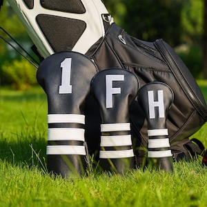 May include: Black golf club head covers with white stripes and large white numbers and letters: 1, F, and H. The covers are in front of a black and white golf bag on a grassy field.