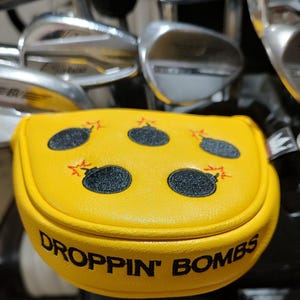 May include: Yellow golf club head cover with a design of black bombs with red explosions. The cover has the words "DROPPIN' BOMBS" in black text. Golf clubs are visible in the background.