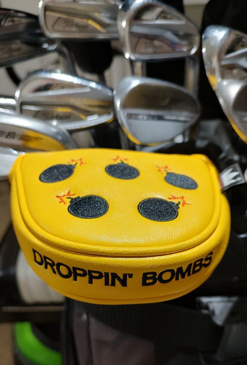 May include: Yellow golf club head cover with a design of black bombs with red explosions. The cover has the words "DROPPIN' BOMBS" in black text. Golf clubs are visible in the background.