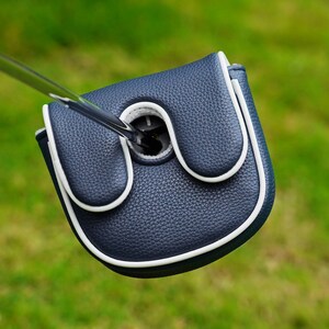 May include: A navy blue golf putter headcover with a textured, pebbled surface and white piping trim. The cover features a U-shaped cutout at the top, allowing the silver metal shaft of the golf club to pass through the center of the protective headcover.