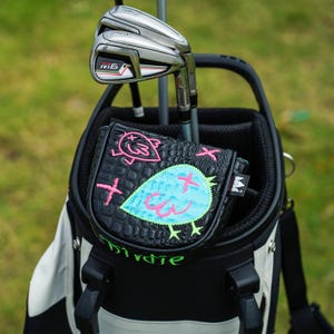 Puede incluir: Una bolsa de golf con palos de golf y una funda de putter negra. La funda del putter tiene un dise&ntilde;o de p&aacute;jaro azul con detalles en rosa y la palabra "Birdie" en verde. La bolsa de golf es negra y blanca.