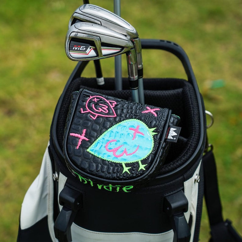 Puede incluir: Una bolsa de golf con palos de golf y una funda de putter negra. La funda del putter tiene un dise&ntilde;o de p&aacute;jaro azul con detalles en rosa y la palabra "Birdie" en verde. La bolsa de golf es negra y blanca.