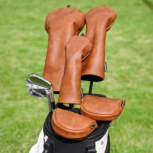 Könnte beinhalten: Eine Golftasche mit einem Satz brauner Golfschlägerhauben aus Leder. Die Hauben sind in verschiedenen Größen und Formen erhältlich, einschließlich Hauben für den Driver, die Fairwayhölzer und den Putter. Die Golfschläger sind in der Tasche sichtbar, mit einem grünen Gras-Hintergrund.