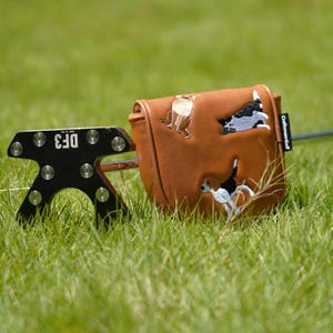 Puede incluir: Un putter de golf negro con el texto "DF3" y una funda de cuero marr&oacute;n para palos de golf bordada con tres dise&ntilde;os de perros. La funda est&aacute; etiquetada como "Craftsman Golf" y descansa en hierba verde.