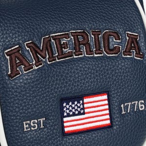 May include: A dark blue textured material featuring the embroidered word AMERICA in brown letters with white outlines. Below the text is an embroidered United States flag with the text EST on the left and 1776 on the right in white stitching.