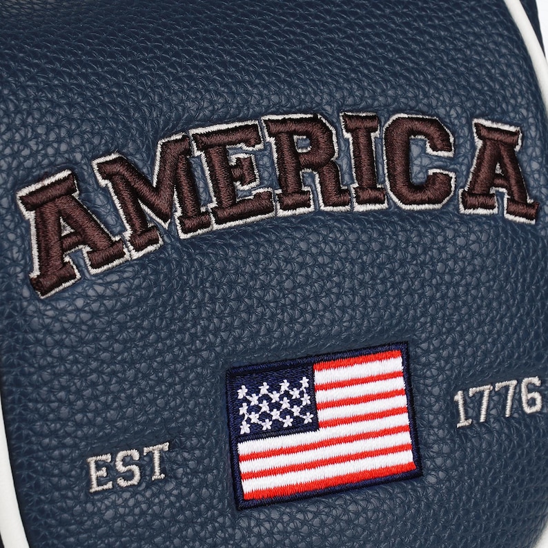 May include: A dark blue textured material featuring the embroidered word AMERICA in brown letters with white outlines. Below the text is an embroidered United States flag with the text EST on the left and 1776 on the right in white stitching.