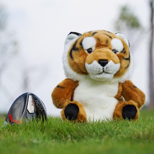 Puede incluir: Una funda para la cabeza del palo de golf con forma de tigre de peluche, con detalles marrones, negros y blancos, est&aacute; sobre hierba verde junto a un palo de golf negro y rojo. El tigre tiene una expresi&oacute;n amigable.