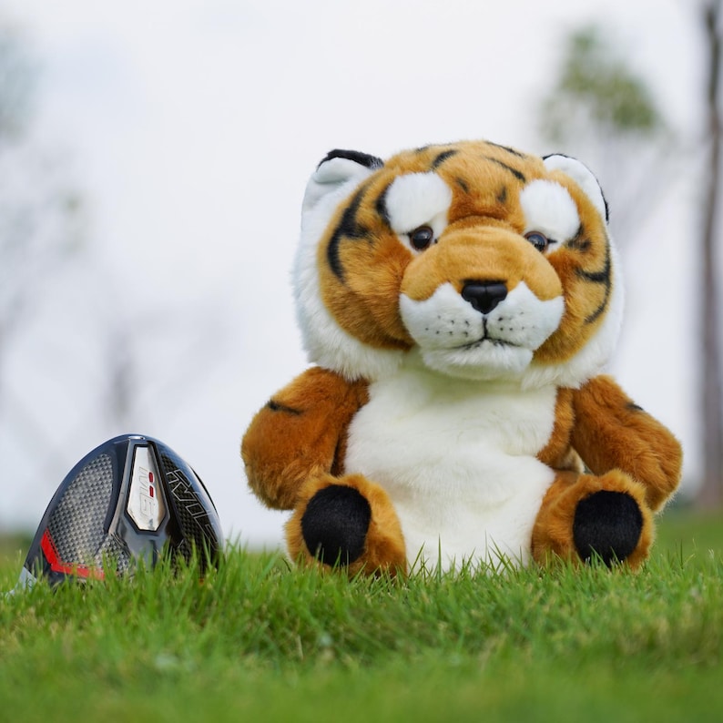 Puede incluir: Una funda para la cabeza del palo de golf con forma de tigre de peluche, con detalles marrones, negros y blancos, est&aacute; sobre hierba verde junto a un palo de golf negro y rojo. El tigre tiene una expresi&oacute;n amigable.