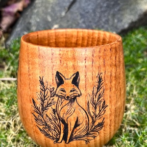 May include: A wooden cup with a fox design burned into the side. The cup is a light brown colour and has a natural wood grain.
