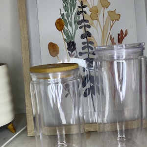 May include: Two clear glass jars with lids, one with a bamboo lid, sit in front of a framed watercolor floral print. The jars are cylindrical and appear to be double-walled. The artwork features muted tones of green, brown, and yellow.