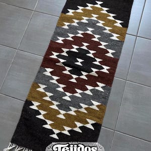 May include: A rectangular, woven rug with a geometric pattern. The rug features a repeating diamond design in shades of black, brown, gray, gold, and white. The ends of the rug have white fringe. The word "Tejidos" is printed on the rug.