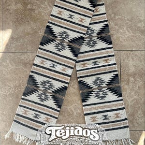 May include: A woven textile runner with a geometric pattern in shades of gray, black, and beige. The runner has a fringed edge and the word "Tejidos" is displayed at the bottom.