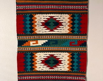 May include: A rectangular, woven wall hanging with a vibrant, geometric design. The textile features a repeating pattern of diamonds, zigzags, and rectangular shapes in red, teal, orange, and white. The bottom of the hanging has the word "Tejidos" in a decorative font.