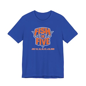 May include: A royal blue short-sleeve t-shirt featuring a graphic design with the orange text FISH IN FIVE centered inside a grey and white triangular emblem. Below the emblem, the text JULY 22, 24, 25, 27, & 29, 2026 is printed in orange.