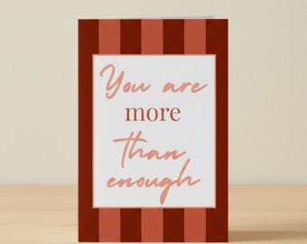 Könnte beinhalten: Eine Grußkarte mit einem rot-weiß gestreiften Rand. Die Karte zeigt die Worte "You are more than enough" in einer kursiven Schriftart. Die Karte steht auf einer hellbraunen Oberfläche.
