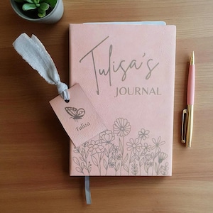 Może przedstawiać: Różowy notatnik z napisem "Tulisa's JOURNAL" i kwiatowym wzorem. Dołączona pasująca zakładka z ilustracją motyla i imieniem "Tulisa". Różowy długopis ze złotymi akcentami spoczywa obok notatnika.