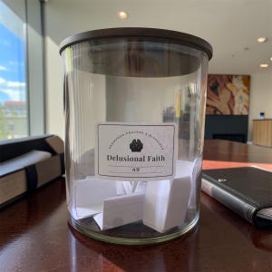May include: A clear glass cylindrical jar with a dark brown lid, containing several folded white paper slips. A rectangular label on the front features the text "ANSWERED PRAYERS & BLESSINGS", a small icon of cupped hands, and "Delusional Faith 49".