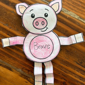 May include: A handmade paper pig craft with pink and white coloring. The pig has a pink circle in the center with the word "Brave" written in cursive. The pig has folded arms and legs, and is set against a wooden background.