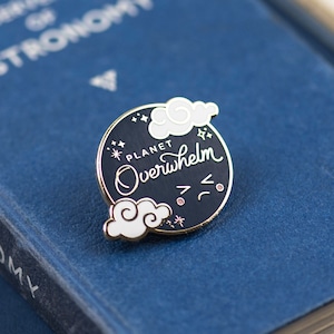 Peut inclure: Une épingle en émail noir avec une lune et des nuages blancs. La lune porte l'inscription "Planet Overwhelm" en lettres blanches cursives. Il y a de petites étoiles blanches autour de la lune et des nuages.