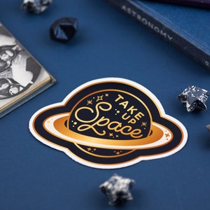 May include: A gold and black sticker featuring a planet design and the text "Take Up Space" in a gold script font.
