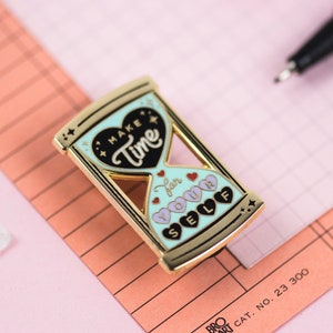 May include: Gold-tone hourglass enamel pin with a blue and green background. The hourglass is filled with black and pink hearts. The text on the pin reads "Make Time for Your Self".