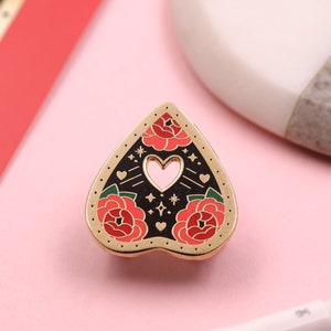 May include: Gold heart-shaped enamel pin with a black background and three red roses with green leaves. The pin has a small heart-shaped cutout in the center and gold glitter accents.