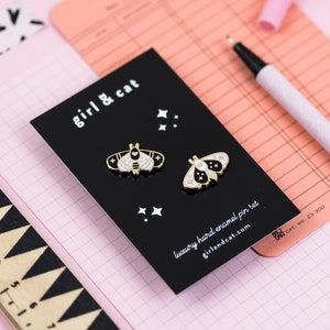 May include: A black card with white text that reads "girl & cat" and "luxury hard enamel pin set" with a website address. Two gold-coloured enamel pins with white and black designs of moths are on the card.