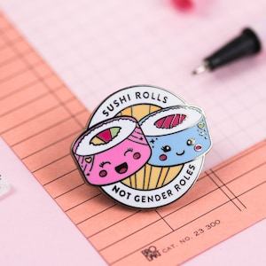 May include: A pink and blue enamel pin featuring two sushi rolls with faces. The pin reads "Sushi Rolls Not Gender Roles".