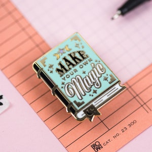 May include: Gold and blue enamel pin shaped like a book with the text "Make Your Own Magic" on the cover. The pin has a black outline and white accents.