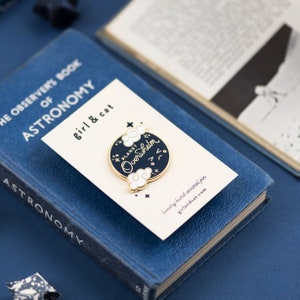 K&ouml;nnte beinhalten: Eine goldene und schwarze Emaille-Anstecknadel mit dem Text "PLANET Overwhelm" in wei&szlig;er Kursivschrift. Die Anstecknadel zeigt einen schwarzen Kreis mit wei&szlig;en Wolken und Sternen. Die Anstecknadel wird auf einer wei&szlig;en Karte mit dem Text "girl & cat" und "luxury hard enamel pin" in Schwarz angezeigt.
