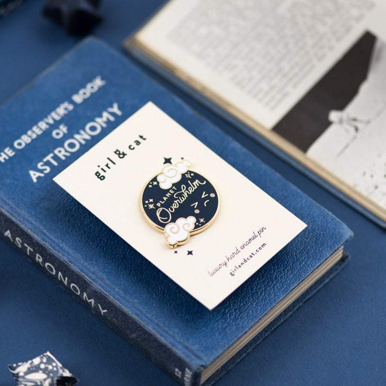 K&ouml;nnte beinhalten: Eine goldene und schwarze Emaille-Anstecknadel mit dem Text "PLANET Overwhelm" in wei&szlig;er Kursivschrift. Die Anstecknadel zeigt einen schwarzen Kreis mit wei&szlig;en Wolken und Sternen. Die Anstecknadel wird auf einer wei&szlig;en Karte mit dem Text "girl & cat" und "luxury hard enamel pin" in Schwarz angezeigt.