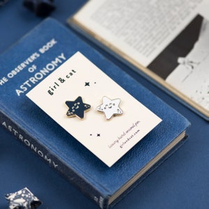May include: Two gold-toned enamel pins shaped like stars. One star is black with white crescent moons and stars. The other star is white with a black smiling face. The pins are on a white card with the text "girl & cat" and "luxury hard enamel pin" and the website "girlandcat.com".