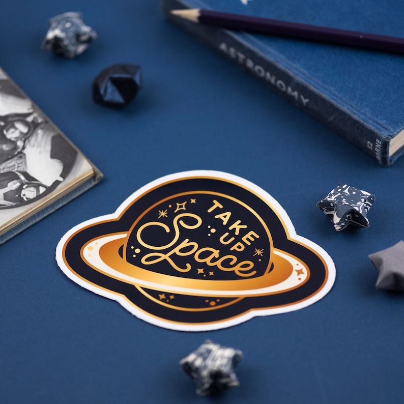 May include: A gold and black sticker featuring a planet design and the text "Take Up Space" in a gold script font.