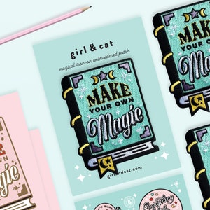May include: A turquoise blue iron-on patch with black and white text that reads "Make Your Own Magic" in a book-shaped design. The patch has a gold star and crescent moon design on the cover.