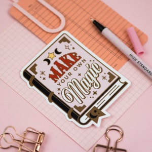 May include: A pink and white sticker with a book design. The book has a black spine and gold accents. The text on the sticker reads "Make Your Own Magic".