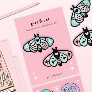 May include: Four iron-on embroidered patches featuring a moth design with pink, blue, and green colors. The patches are on a pink background with the text "girl & cat" and "magical iron-on embroidered patch".