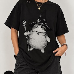May include: A black short-sleeved t-shirt featuring a graphic print of a fluffy tabby cat wearing a pointed witch hat decorated with stars and moons. The cat holds a tall, slender staff with a spherical top and wears a collar with a small bell.