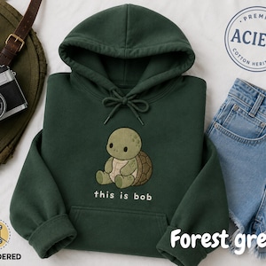 Puede incluir: Una sudadera con capucha de color verde bosque que presenta un gráfico bordado de una pequeña tortuga de color verde claro sentada. Debajo de la tortuga, el texto "this is bob" aparece en letras blancas. Incluye bolsillo frontal y cordones.