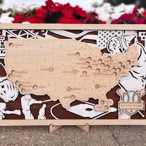May include: A rectangular wooden wall art piece featuring a laser-cut map of the United States with circular cutouts for tracking baseball stadiums. The design includes baseball-themed silhouettes, a flag, and a plaque labeled Ballpark Tracker.