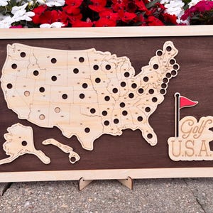May include: A rectangular wooden display board featuring a light-colored map of the United States with circular cutouts for each state. A red golf flag icon and the text Golf U.S.A. are positioned on the right side against a dark brown background.